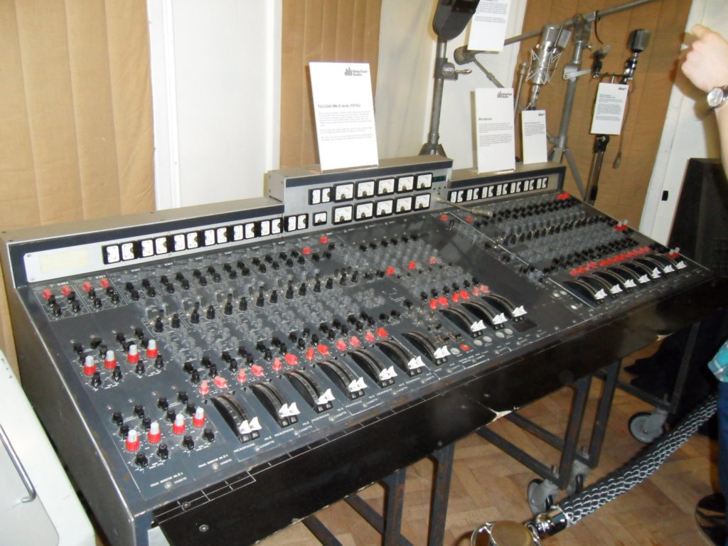 A 1970s-era EMI TG12345 Mk.II recording console displayed in a museum with vintage microphones. Photo by Clusternote used under Creative Commons Attribution license: https://creativecommons.org/licenses/by/2.0/deed.en